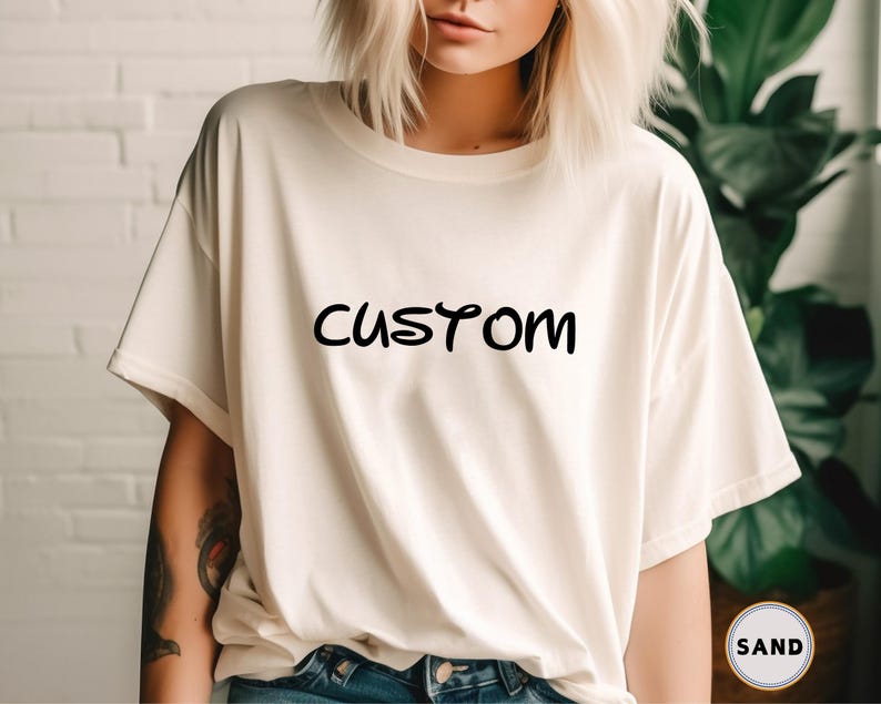 以下が含まれることがあります： 前面に黒い文字で「CUSTOM」とプリントされた白いTシャツです。