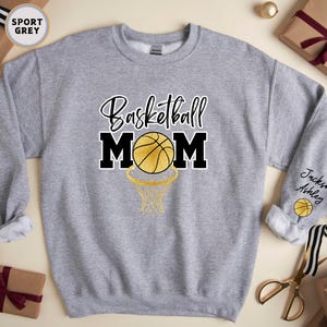 May include: A grey sweatshirt with the text "Basketball Mom" in black and gold lettering. A basketball with gold accents is inside a hoop. The sleeve has the text "Jackson Ashley" and a basketball.