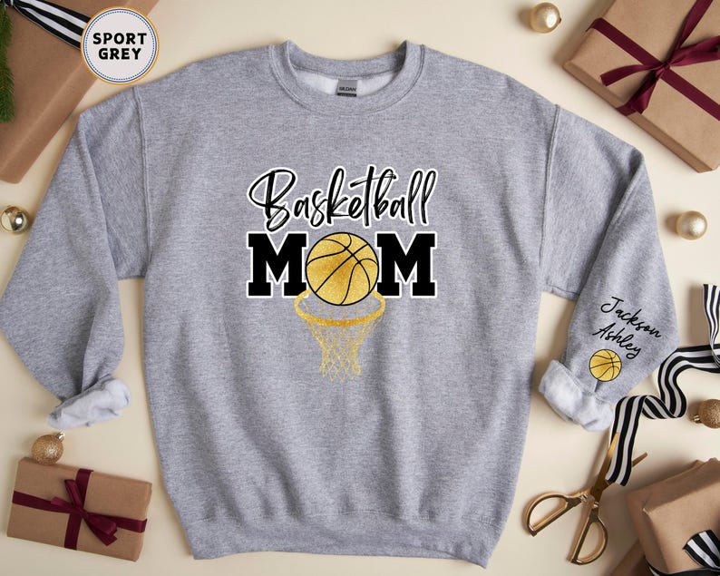 May include: A grey sweatshirt with the text "Basketball Mom" in black and gold lettering. A basketball with gold accents is inside a hoop. The sleeve has the text "Jackson Ashley" and a basketball.