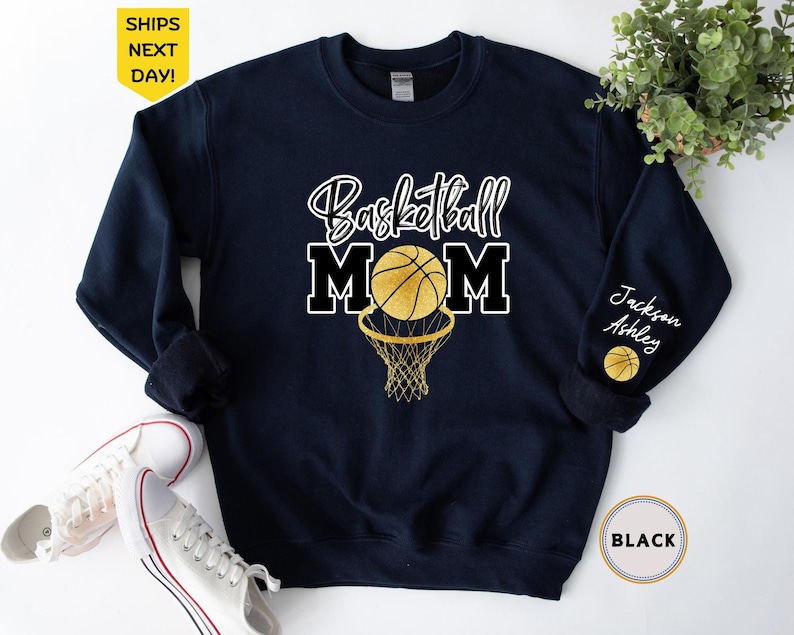 May include: A navy blue sweatshirt with the text "Basketball Mom" in black and white lettering. A gold basketball with black lines is going through a gold basketball hoop. The text "Jackson Ashley" is on the sleeve of the sweatshirt.