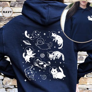 Peut inclure: Un sweat &agrave; capuche bleu marine avec un motif blanc de chats dans l'espace. Le motif repr&eacute;sente des chats en combinaison spatiale, des plan&egrave;tes et des &eacute;toiles. Le sweat &agrave; capuche a une capuche avec cordon de serrage et une poche kangourou.