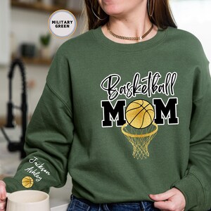 May include: A green sweatshirt with the text "Basketball Mom" in black and white lettering. A basketball with gold accents is inside a black hoop. The sweatshirt has a personalised name on the sleeve.