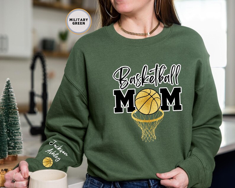 May include: A green sweatshirt with the text "Basketball Mom" in black and white lettering. A basketball with gold accents is inside a black hoop. The sweatshirt has a personalised name on the sleeve.