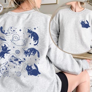 Peut inclure: Un sweat-shirt gris avec un imprim&eacute; de chats dans l'espace. Les chats portent des casques d'astronautes et sont entour&eacute;s d'&eacute;toiles et de plan&egrave;tes. Le texte "SPORT GREY" est imprim&eacute; dans le coin sup&eacute;rieur gauche du sweat-shirt.