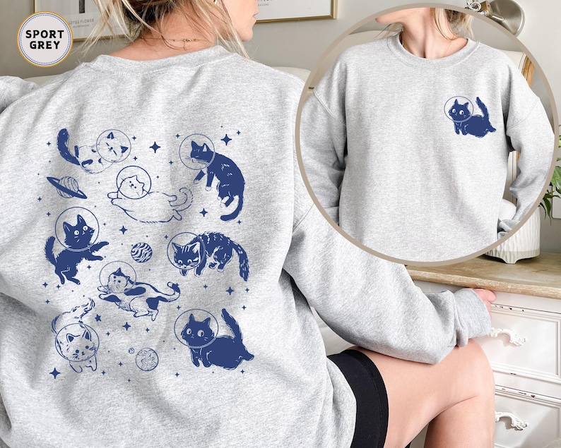 Peut inclure: Un sweat-shirt gris avec un imprim&eacute; de chats dans l'espace. Les chats portent des casques d'astronautes et sont entour&eacute;s d'&eacute;toiles et de plan&egrave;tes. Le texte "SPORT GREY" est imprim&eacute; dans le coin sup&eacute;rieur gauche du sweat-shirt.