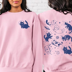 Peut inclure: Un sweat-shirt rose p&acirc;le avec un motif de chats dans l'espace. Les chats portent des casques d'astronautes et flottent dans l'espace. Le texte "Light Pink" est imprim&eacute; dans le coin sup&eacute;rieur gauche de l'image.