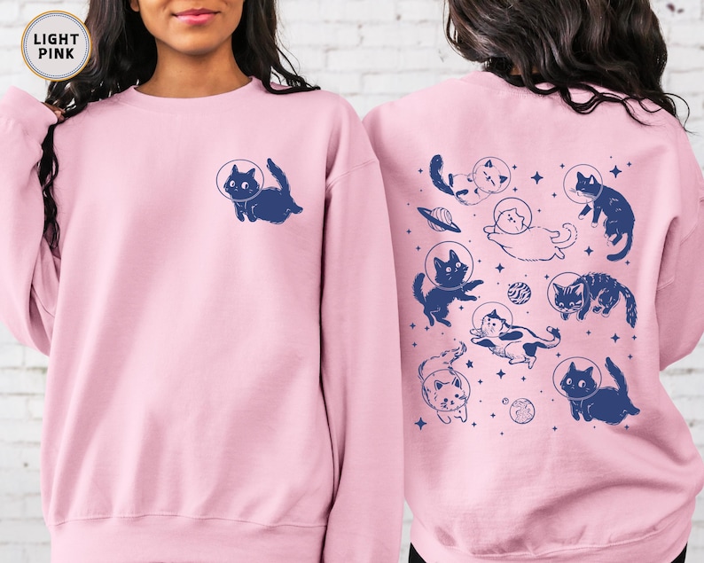 Peut inclure: Un sweat-shirt rose p&acirc;le avec un motif de chats dans l'espace. Les chats portent des casques d'astronautes et flottent dans l'espace. Le texte "Light Pink" est imprim&eacute; dans le coin sup&eacute;rieur gauche de l'image.