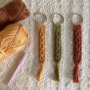 May include: An overhead shot displays three handmade macrame keychains in shades of gold, olive green, and rust, each with a tassel. Two skeins of yarn and a crochet hook are also visible. The keychains are on a white textured surface.