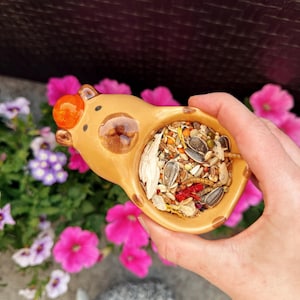 May include: A tan ceramic hamster-shaped bowl filled with a mix of seeds and grains. The bowl has a round orange nose and is held in front of a backdrop of pink and purple flowers.