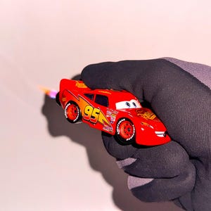 May include: A red toy car, featuring the number 95 and lightning bolt graphics, is held in a black glove. The car has a flame effect coming from the back. The car is a replica of Lightning McQueen from the film Cars.