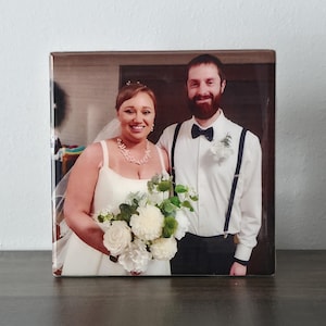 May include: A square photo print featuring a smiling couple on their wedding day. The bride wears a white wedding dress and holds a bouquet of white and green flowers. The groom wears a white shirt, black bow tie, and suspenders.