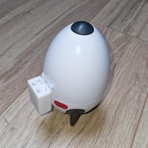 May include: A white, egg-shaped object with a dark grey top and a small, rectangular white attachment. It has a red accent and a black, T-shaped base. The object is on a wooden surface.