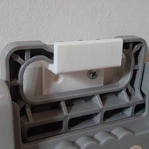 May include: A close-up of a white plastic wall mount with a screw. The mount is attached to a grey plastic object with a handle.