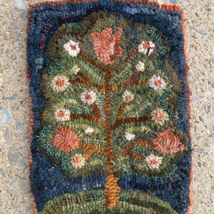 May include: A hand-stitched rug depicting a stylized tree with pink and white flowers. The rug has a dark blue border and is made with a variety of wool colors.