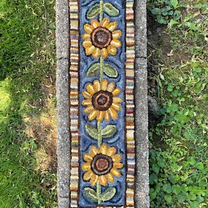 May include: Hand-hooked sunflower rug with three large yellow sunflowers on a blue background. The rug has a rectangular shape with a border of various colors. The text "Hand and Wing Studio" is at the bottom of the rug.