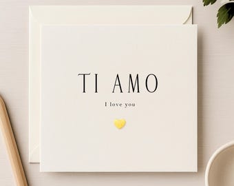 May include: A cream-colored greeting card with the words "TI AMO" and "I love you" printed in black. A small, gold heart is below the text. The card is inside a matching envelope.