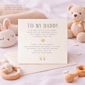 May include: A cream-colored card with the message "TO MY DADDY" and a baby's message, finished with gold leaf. The card is surrounded by baby toys, including a teddy bear and a bunny.