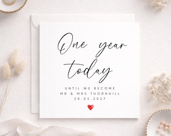 May include: A white card with the text "One year today" in black script. Below, it reads "Until we become Mr & Mrs Thornhill 28.05.2027" with a red heart. The card is on a white surface with wedding rings and decorative elements.
