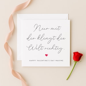 May include: A white Valentine's Day card with black script and a small pink heart. The card is next to a red rose and a pink ribbon. The text on the card reads "Nur mit dir klingt die Welt richtig."