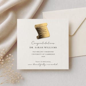 May include: A square white card with a gold foil scroll icon at the top. The text reads: Congratulations, DR. SARAH WILLIAMS, PhD ORGANIC CHEMISTRY, UNIVERSITY OF CAMBRIDGE, 2026, Years of dedication, now beautifully rewarded. Includes an envelope.