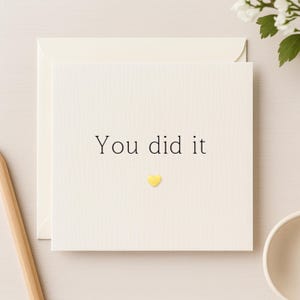 Könnte beinhalten: Cremefarbene Grußkarte mit den Worten "You did it" in schwarzer Schrift. Unter dem Text befindet sich ein kleines goldenes Herz. Die Karte befindet sich in einem cremefarbenen Umschlag.