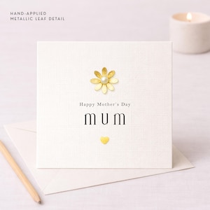 Può includere: Un biglietto color crema per la festa della mamma con un abbellimento a margherita dorata e un centro di perle. Il biglietto recita "Happy Mother's Day" e "MUM" in testo nero, con un piccolo cuore dorato sotto. Le parole "HAND-APPLIED METALLIC LEAF DETAIL" sono in alto.