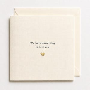 May include: A square cream-colored greeting card with a textured finish. The text "We have something to tell you" is printed in black ink in the center, positioned above a small, textured gold-colored heart icon. An envelope is tucked behind the card.