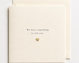 May include: A square cream-coloured greeting card with a textured finish. The text "We have something to tell you" is printed in black ink in the centre, positioned above a small, textured gold-coloured heart icon. An envelope is tucked behind the card.