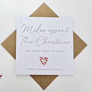 Puede incluir: Una tarjeta de Navidad blanca con el mensaje "Miles apart This Christmas But never apart in heart" en escritura negra. La tarjeta está sobre un sobre marrón, con dos bastones de caramelo y un bastón de caramelo en forma de corazón en el centro. La tarjeta dice "Merry Christmas Chloe".