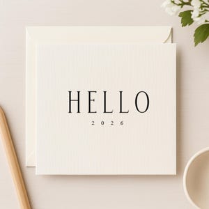 Puede incluir: Una tarjeta color crema con la palabra "HELLO" en una fuente negra y elegante, y el número "2026" debajo. La tarjeta está dentro de un sobre a juego. Se ven un lápiz de madera y un cuenco blanco pequeño.