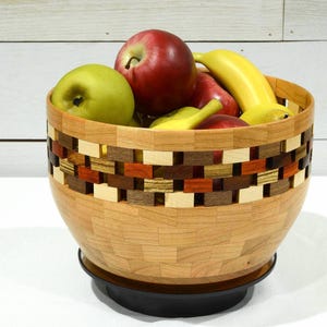 May include: A wooden fruit bowl with a geometric pattern in shades of brown, red, and yellow. The bowl is filled with green apples, red apples, and yellow bananas.