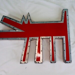 May include: A large, stylized metal dog sculpture with a red body and a silver-colored, riveted frame. The dog's form is geometric, with a pointed head and tail, and rectangular legs. The sculpture is likely for outdoor or indoor decor.