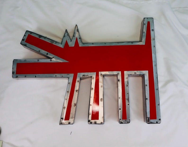 May include: A large, stylized metal dog sculpture with a red body and a silver-colored, riveted frame. The dog's form is geometric, with a pointed head and tail, and rectangular legs. The sculpture is likely for outdoor or indoor decor.