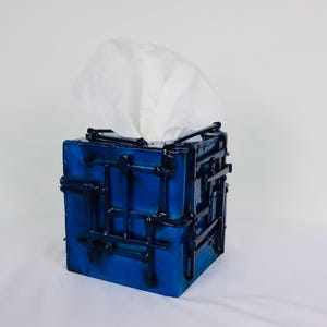 May include: A square tissue box cover featuring a vibrant blue finish with a raised, grid-like pattern of dark black metal bars welded across the exterior surfaces. A single white tissue is pulled through the top opening of the container.