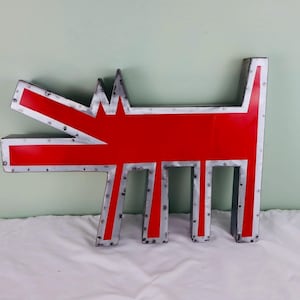 May include: A large, stylized metal dog sculpture. The dog is primarily red with silver-colored trim and rivets. The dog's mouth is open, and it has a jagged crown-like shape on its head. The sculpture is on a white surface.