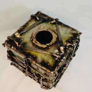 May include: A square, handcrafted metal box with a central circular opening. The box features a textured, layered design with a mix of green, gold, and black tones. The metalwork appears to be welded and has a rustic, industrial aesthetic.