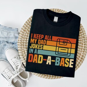 May include: Black t-shirt with the text "I KEEP ALL MY DAD JOKES IN A DAD-A-BASE" in orange, red, yellow, and teal. The design includes a retro-style floppy disk graphic. The shirt is on a woven mat with denim shorts and white sneakers.