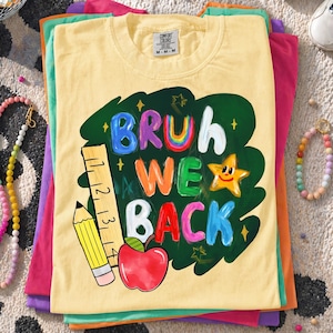 May include: A pale yellow short-sleeve t-shirt featuring a dark green chalkboard-style graphic with the text "BRUH WE BACK" in colorful, hand-painted style letters. The design includes a yellow pencil, a ruler, a red apple, and a smiling star.
