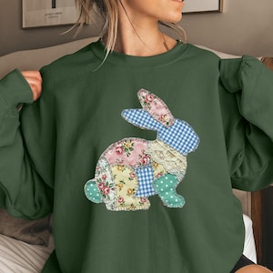 May include: A dark green sweatshirt with a patchwork bunny motif. The bunny is made from assorted fabrics, including floral patterns, gingham, and polka dots. The design is positioned centrally on the front of the sweatshirt.