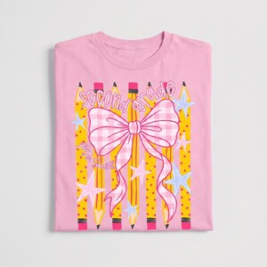 May include: A light pink t-shirt featuring a graphic print of yellow pencils with polka dots, a large pink and white gingham bow, and light blue stars. The text "second grade" is written in a cursive font, and "Mrs. Smith" is printed below.