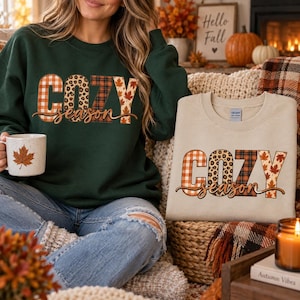 May include: A dark green crewneck sweatshirt and a sand-colored crewneck sweatshirt, both featuring the text "COZY season" in a multi-patterned font with orange plaid, leopard print, dark plaid, and autumn leaf designs on a white mug.