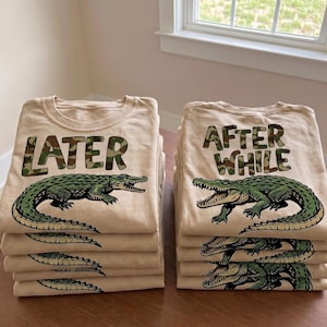 Peut inclure: Piles de t-shirts beiges avec des illustrations d'alligators verts. Une pile affiche le texte "LATER" en lettres camouflage, et l'autre lit "AFTER WHILE" en lettres camouflage. Les t-shirts sont empilés sur une surface de couleur claire.