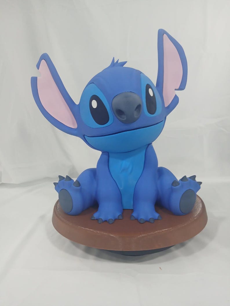 May include: A blue figurine of the character Stitch from the animated film Lilo & Stitch. The figurine has large ears with pink inner ears, large black eyes, and a dark blue nose. It is sitting on a brown circular base.