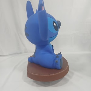 May include: A blue Stitch figurine, seated on a brown base. The figurine features large ears, a round head, and a detailed facial expression. The figure is a collectible item.