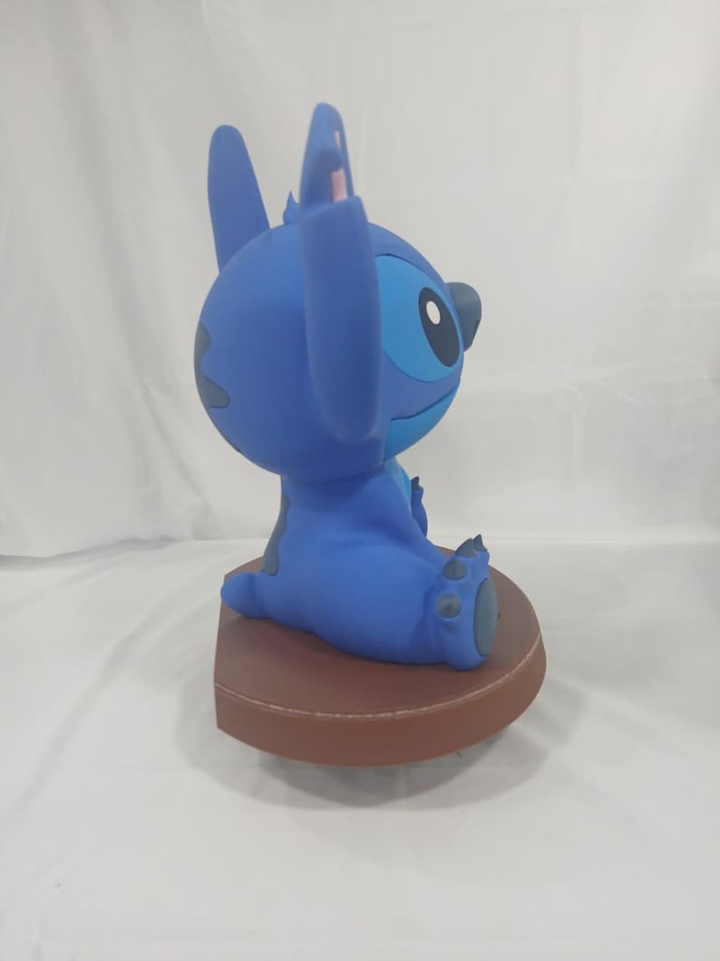 May include: A blue Stitch figurine, seated on a brown base. The figurine features large ears, a round head, and a detailed facial expression. The figure is a collectible item.