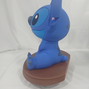 May include: A blue Stitch figurine, seated on a brown base. The figurine features large ears, a round head, and a playful expression. The figure is a popular character from a well-known animated film.