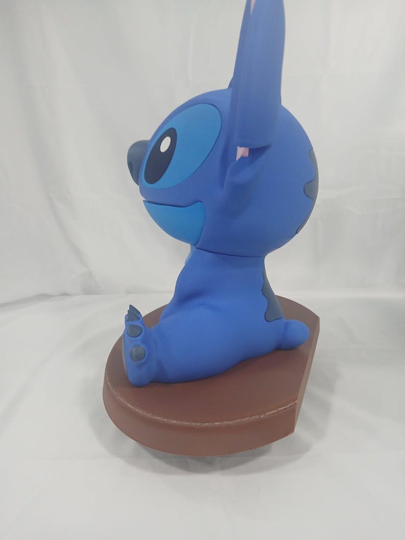 May include: A blue Stitch figurine, seated on a brown base. The figurine features large ears, a round head, and a playful expression. The figure is a popular character from a well-known animated film.