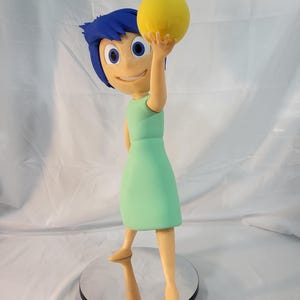 May include: A figurine of the character Joy from the animated film Inside Out. Joy is depicted with blue hair, a mint green dress, and is holding a yellow orb. The figurine is standing on a round, mirrored base.