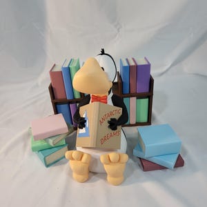 May include: A whimsical penguin figurine with a large yellow beak and a red bow tie, holding a book titled "Antarctic Dreams." The penguin is surrounded by miniature books in various colors, with a bookshelf in the background.