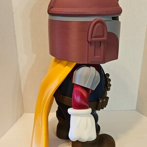 Op de afbeelding: Een rood, wit en grijs 3D-geprint actionfiguur van een personage met een helm, een gele cape en een bruine riem met een bandoleer. Het figuur staat op een wit oppervlak.